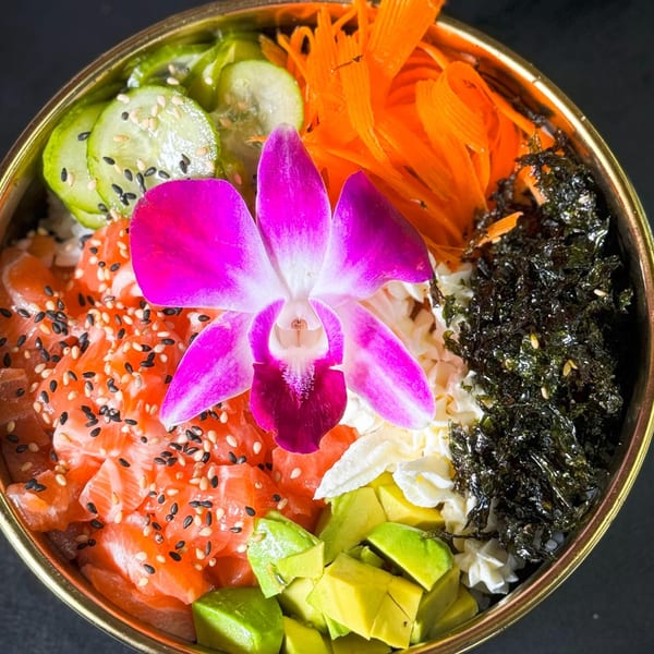 A vibrant poke bowl with salmon, avocado, cucumber, carrots, seaweed, and a pink orchid.