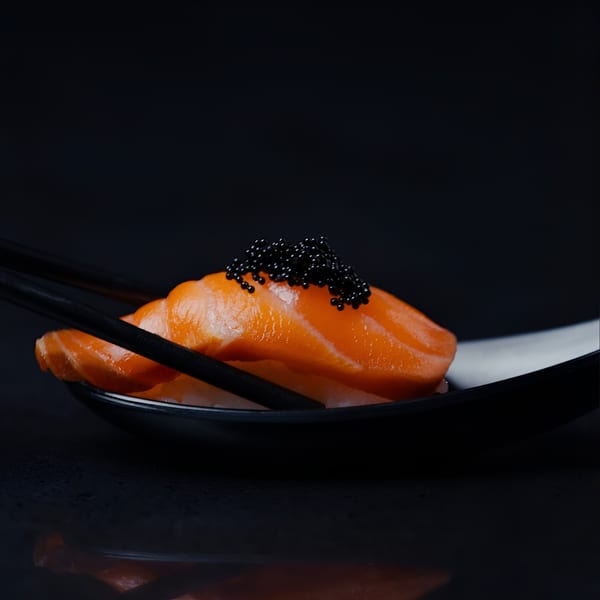 Salmon nigiri with black caviar on a dark plate, held by black chopsticks.
